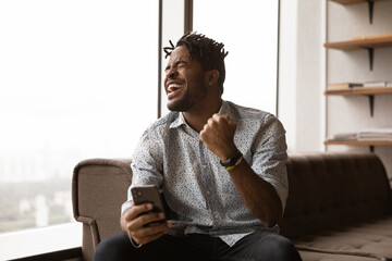 Overjoyed black male hipster blogger receive perfect commercial offer pleasant comment at social network. Excited young african man watch lottery result on smartphone screen celebrate winning jackpot