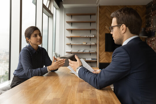 Young man professional lawyer counsel give advice propose decision to serious indian woman customer at private office. Young mixed race female visit male insurance broker talk about policy contract
