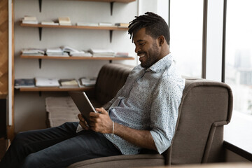 Young black man sit on sofa at modern office wait for job interview meeting appointment spend time for reading electronic book on tablet pc. Relaxed african guy hipster chat online using digital pad