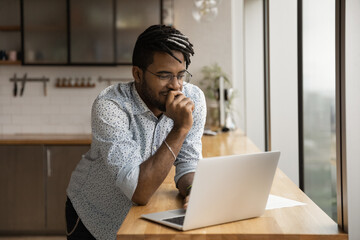 Smiling black guy freelancer stand at kitchen desk read good email message on laptop screen think on advantages proposed by customer. Young afro man hipster chat with friends online from home using pc