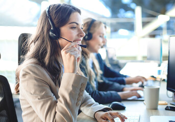 Female customer support operator with headset and smiling, with collegues at background.