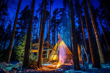 Winter teepee. Tipi in the woods at night. The lighted tent is surrounded by pine trees. Teepee against a beautiful sky. Ethnic housing in the winter forest. © Grispb