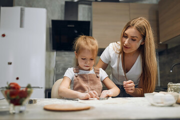 Happy little chef, flour, knead the dough. The girl cook rolls out the dough with a rolling pin. Home cooking. Mom is doing her daughter's hair. kitchen