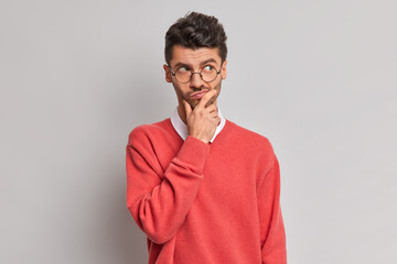Photo of thoughtful handsome adult European man holds chin and looks pensively away tries to solve problem wears round glasses and red jumper isolated over grey background. Let me think about it