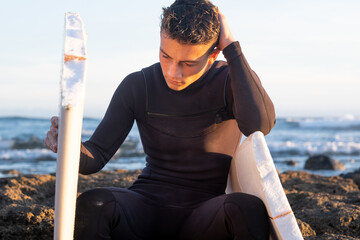 Young surfer sitting on the rocks looks thoughtfully at the broken board in half. Handsome guy with...