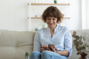 Smiling young Caucasian woman sit rest on couch at home look at cellphone screen browse wireless internet on gadget. Happy female relax on sofa in living room text message online on smartphone.