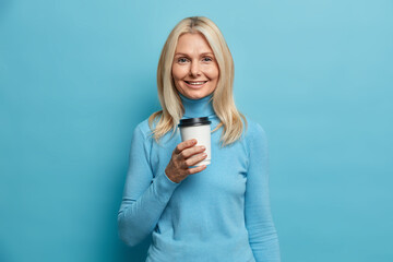 Portrait of good looking adult European woman holds disposable cup of coffee dressed in casual...