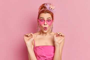 Surprised redhead young European woman wears trendy sunglasses raises hands stares at camera and says omg dressed in festive dress isolated over pink background. People style fashion concept