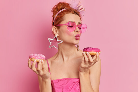Photo of fashionable redhead glamour European woman keeps lips folded holds two appetizing doughnuts wants to eat sweet dessert wears earrings sunglasses and dress isolated over pink background