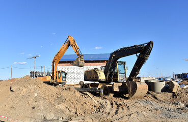 Obraz premium Excavator on earthworks at construction site. Backhoe on foundation work and road construction. Tower cranes in action on blue sky background. Heavy machinery and construction equipment