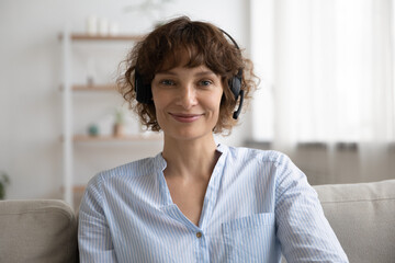 Portrait of smiling young Caucasian woman in headphones sit on sofa at home talk on video call. Happy female in earphones have webcam digital conference or online virtual communication with client.