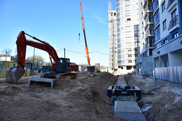 Excavator on earthworks for laying heating pipes and stormwater in trench. Installing concrete wells, сhambers, manholes near new multi-storey residential building. Tower cranes at construction site