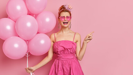 Obraz na płótnie Canvas Crying dejected redhead young woman feels unhappy indicates aside on copy space demonstrates something wears fashionable pink dress and shades holds bunch of balloons isolated over rosy wall