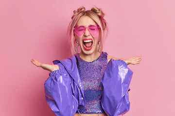 Portrait of blonde woman exclaims loudly spreads palms dressed in fashionable purple outfit wears pink trendy sunglasses isolated over rosy background. People negative emotions style concept