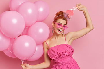 Obraz na płótnie Canvas Half length shot of cheerful European woman has upbeat mood dances carefree with balloons and sweet candy poses against rosy background being on party. People holidays and celebration concept