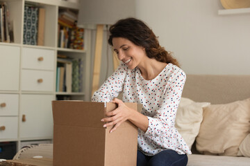 Fototapeta na wymiar Smiling young Caucasian woman sit on sofa at home unbox package with internet order. Happy excited millennial female client or buyer unpack box shopping online. Good delivery service concept.