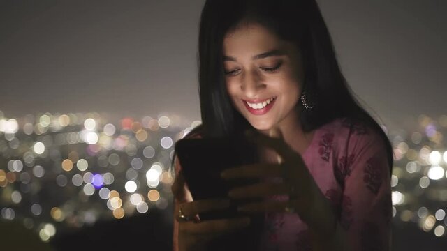 close up shot of a cheerful young Indian female sitting on a terrace of building having a virtual conversation through messaging using a mobile phone against the illuminated cityscape at night - Powered by Adobe