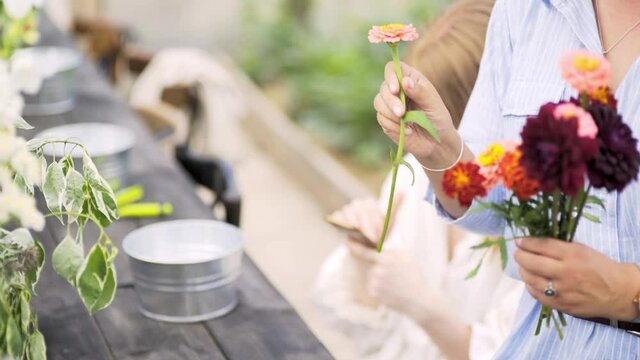 Unrecognizable woman in light blue dress puts bouquets with colorful flowers in vases on long wooden table preparing for masterclass in greenhouse or decorate for event celebration