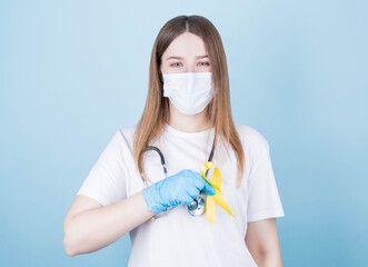 a brunette girl, a doctor in a medical mask with a stethoscope around her neck, in a white T-shirt, smiles and holds a yellow ribbon near her chest, a symbol of health on a blue background