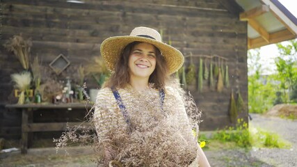 Portrait woman in yellow stripe shirt and straw hat enjoying fresh bouquet with gentle windflowers. Lady smiling and looking at camera at background summerhouse wall with dried grass 