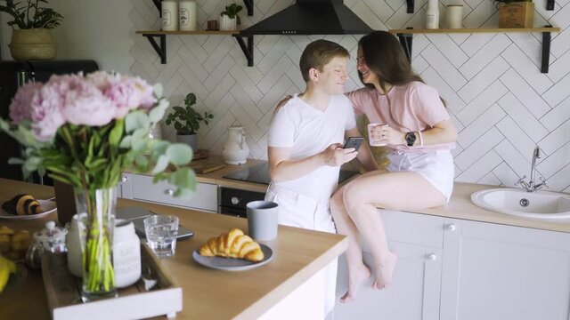 Happy young couple in casual clothes uses smartphone at modern kitchen. Man shows the screen to a cheerful woman sitting on wooden counter while have breakfast with coffee and croissant
