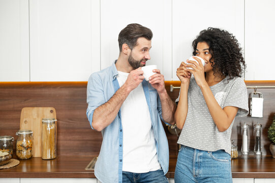 Interracial couple drinks morning coffee in the kitchen at home. An African woman and a caucasian guy enjoy hot drinks at home