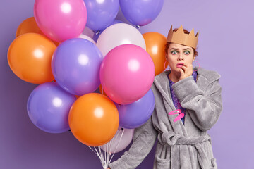 Fototapeta na wymiar Shocked woman celebrates anniversary gets congratulations and undexpected gifts dressed in casual dressing gown holds inflated colorful balloons isolated over purple background. Holiday concept