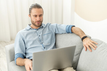 Handsome guy spends time in networks, a young man lying on the comfortable couch and using a laptop, watching movies, working remotely from home