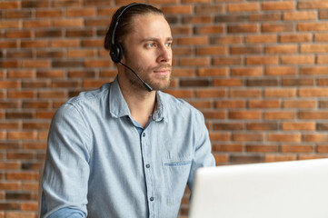 Thoughtful guy wearing a headset looks away, a pensive call center employee sits at the desk in...