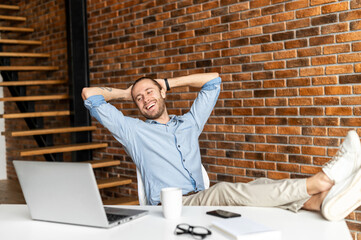 Cheerful young businessman leaned back in the chair, holds his hands behind his head and looks at...