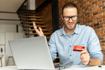 A puzzled guy sits at the desk with a laptop, holds credit card in hand and looks at it with incomprehension, a man could not complete the transaction online, not enough money on the card