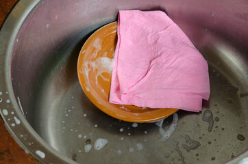 Pink viscose dishcloth and brown plate in the metal kitchen sink with foam.