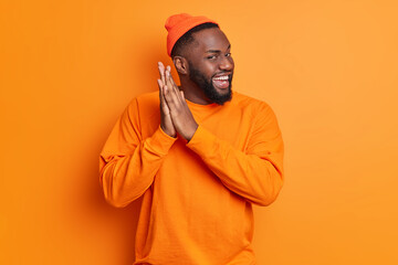 Positive dark skinned man rubs palms and smiles gladfully has intention to do something wears hat and sweater isolated over vivid orange background. Bearded African American male feels glad. - Powered by Adobe
