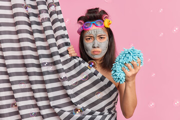 Displeased gloomy Asian woman with dark hair makes hairstyle applies facial clay mask for skin rejuvenation holds shower sponge has angry mood poses behind curtain isolated on pink studio wall