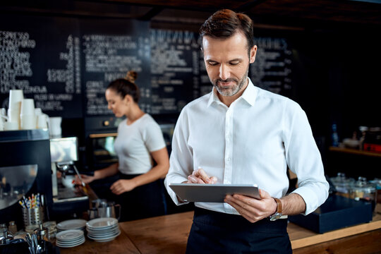 Restaurant manager standing with digital tablet with employee in background preparing coffee