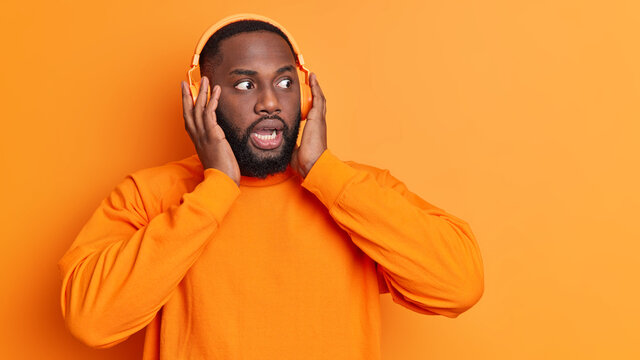 Dark skinned bearded man stares bugged eyes keeps hands on stereo headphones listens music reacts on something dressed in casual jumper isolated over vivid prange background with copy space.