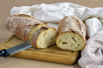 Sliced fresh bread loaf on wooden cutting board. Baked bread, knife and dish towel lies on table on craft paper, home baked loaf. Natural color. Healthy eating. Close-up. Selective focus.