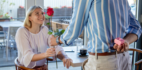 Young romantic couple on a date in a cafe or restaurant. Boyfriend surprises his girlfriend with a red rose and an engagement ring behind his back. Selective focus on the hand behind the back.