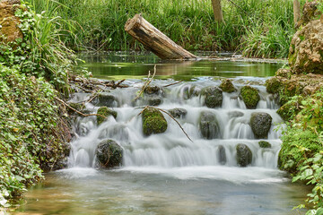 Foto del nacimiento del río Huéznar con una mini cascada llena de piedras con verdina y un tronco.