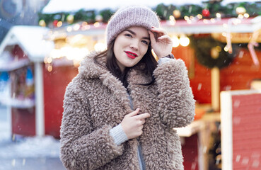 Young girl in beige fur coat and cap posing next to the christmas fair