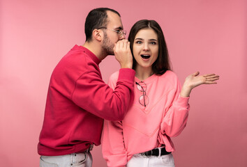 Photo of sweet couple, man whispers some secrets or sales to his girlfriend and she is surprised, isolated over pink background