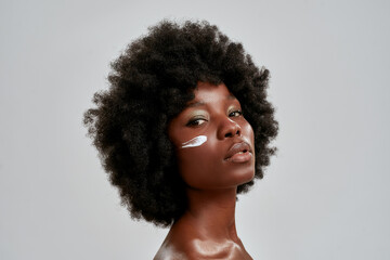 Beauty portrait of young naked african american woman with afro hair looking at camera while posing with cream applied on her face isolated over gray background - obrazy, fototapety, plakaty