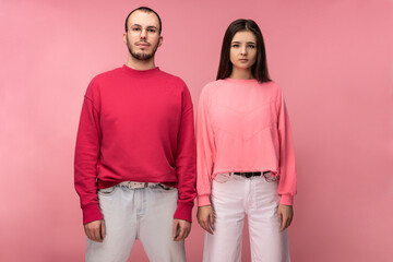 Photo of attractive man wih beard in red clothing and woman in pink stand with serious face, isolated over pink background