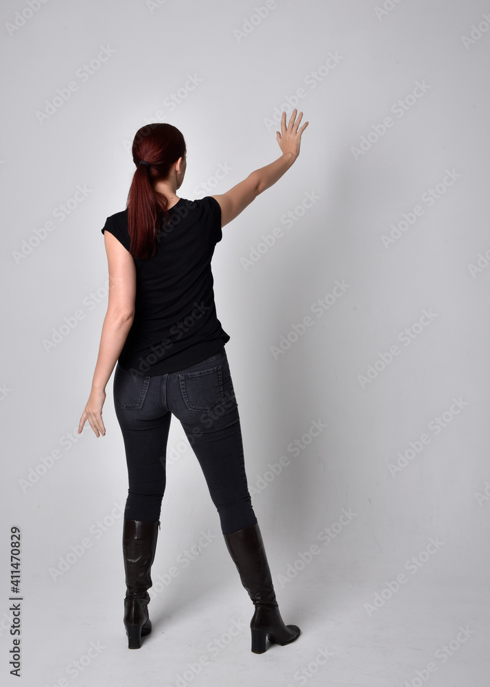 Sticker Full length portrait of woman with red hair in a ponytail, wearing black t-shirt and denim pants. Standing pose, hands reaching out with back to the camera the camera, against a  studio background.