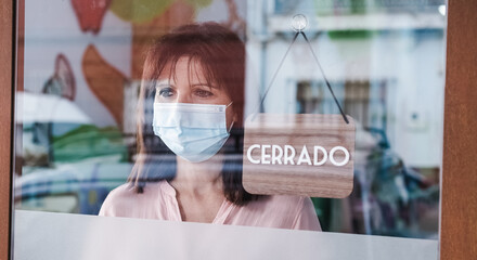 sad woman with face mask changing open to closed sign in spanish on window for corona virus