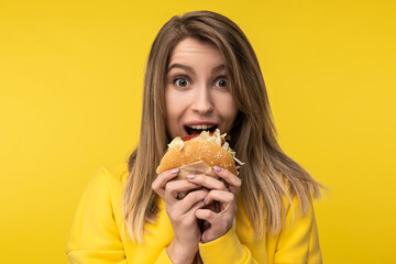 Photo of attractive lady poses in a happy way tries to bite burger. Wears casual yellow hoody, isolated yellow color background