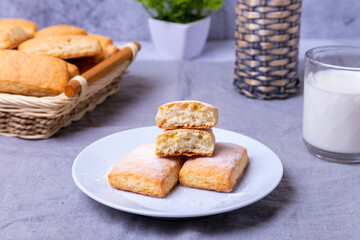 Cookies with powdered sugar on a white plate. In the background a basket of cookies and a cup of milk. Close-up.