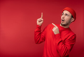 Naklejka premium Photo of attractive man with beard in glasses and red clothing. Shocked and puzzled male points up, isolated over red background