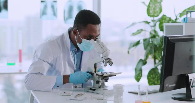 Young african medical worker in face mask examining vaccine liquid with microscope wearing gloves. Development of vaccination.