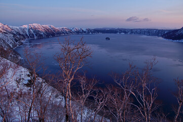 冬の夜明けの山間の湖の薄く凍った湖面と周囲の山。日本の北海道の阿寒摩周国立公園の摩周湖。